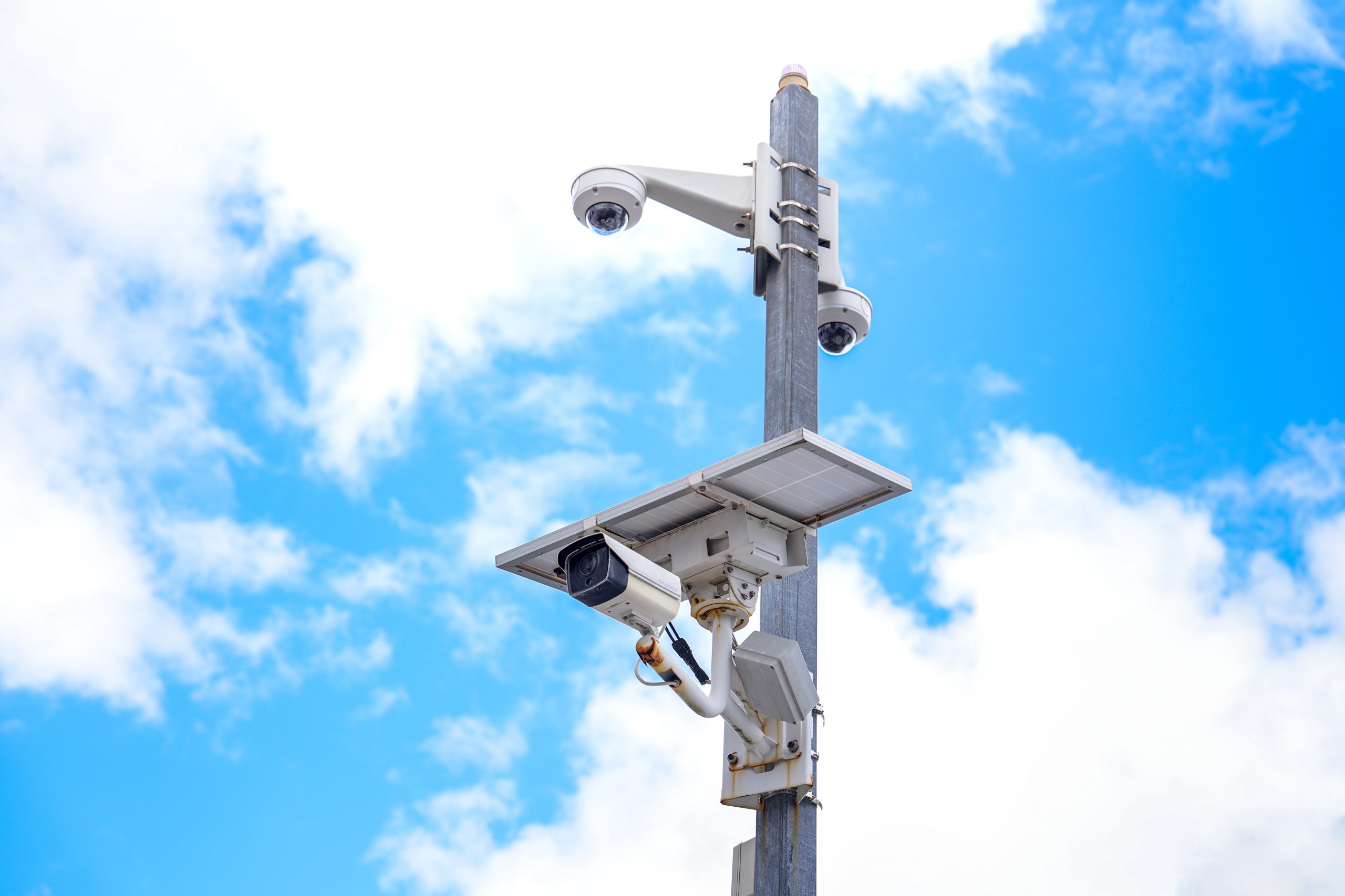This image features a modern surveillance camera mounted on a pole, set against a clear blue sky with fluffy clouds, emphasizing technology and outdoor security solutions.